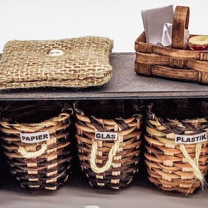 Può includere: Uno scaffale in legno con tre cesti intrecciati etichettati "Papier", "Glas" e "Plastik". Un piccolo cuscino di juta si trova sullo scaffale superiore, e un cestino in vimini in miniatura con un tovagliolo di carta bianco e una mela rossa si trova sullo scaffale superiore.