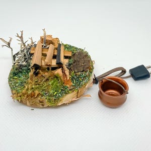 Pode incluir: Um diorama em miniatura de uma cena de fogueira. A cena inclui uma pequena panela de cobre, uma base de madeira com musgo e toras em miniatura e um plugue com um cabo. O diorama foi projetado para ser conectado.