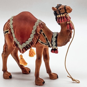 Op de afbeelding: Een bruin decoratief kameelbeeldje met een rood zadel en gouden afwerking. De kameel heeft een zwarte en gouden teugel en decoratieve kwastjes. Een touw is aan de kop van de kameel bevestigd.