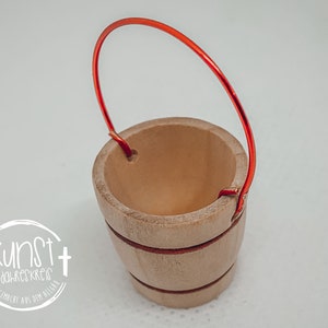 Puede incluir: Un peque&ntilde;o cubo de madera con un asa de alambre rojo. El cubo tiene un acabado de madera natural y est&aacute; decorado con una fina franja marr&oacute;n alrededor del medio.