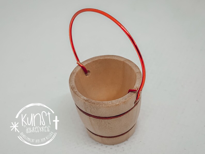 Puede incluir: Un peque&ntilde;o cubo de madera con un asa de alambre rojo. El cubo tiene un acabado de madera natural y est&aacute; decorado con una fina franja marr&oacute;n alrededor del medio.