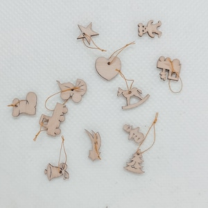 May include: A set of 12 wooden Christmas ornaments with a variety of shapes, including a star, a heart, a rocking horse, a truck, a bell, a snowman, a tree, and a few other abstract shapes. The ornaments are all made of natural wood and have a rustic, handmade look.
