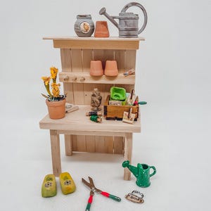 Könnte beinhalten: Miniatur-Gartentisch aus Holz mit Gartengeräten und Zubehör. Enthält eine Gießkanne, Holzschuhe und einen kleinen Blumentopf mit gelben Blumen. Die Werkbank hat ein Regal mit kleinen Töpfen und eine Gießkanne.
