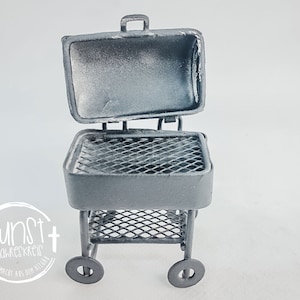 Può includere: Un barbecue in miniatura in metallo grigio con coperchio aperto, griglia e ripiano inferiore. Il barbecue ha due ruote e una maniglia in alto. Il testo "kunst jahreskreis handgemacht aus dem allgäu" è in un cerchio bianco in basso a sinistra.