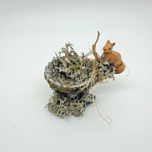 May include: A miniature woodland scene with a wooden squirrel on a moss-covered bowl. The bowl contains dried plants and pink berries. The base is covered in green moss, creating a naturalistic aesthetic. The scene is a detailed, decorative piece.