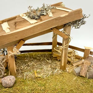 Op de afbeelding: Een miniatuur houten stal met een verweerde uitstraling. Het dak is gemaakt van ruwe planken en de constructie omvat houten palen, een met hooi bedekte vloer en decoratief mos en twijgen. Een kleine steen staat aan de basis.