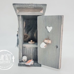 Könnte beinhalten: Ein Miniatur-Holzhäuschen in Grau mit offener Tür. Im Inneren befinden sich eine kleine Holztoilette und eine Rolle Toilettenpapier. Die Tür hat einen herzförmigen Ausschnitt und ein kleines Papier. Der Text "kunst jahreskreis handgemacht aus dem allgäu" ist auf der Vorderseite.