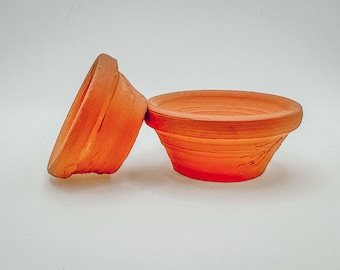 Può includere: Due vasi in terracotta, uno è una piccola ciotola e l'altro è un vaso più alto e stretto. Entrambi sono di colore marrone rossiccio.