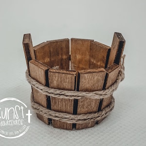 Könnte beinhalten: Ein kleiner Holz-Eimer mit einem Seilhenkel. Der Eimer besteht aus dünnen Holzlatten und ist mit einem Seil zusammengebunden.