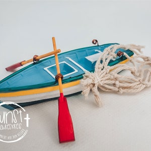 Puede incluir: Un pequeño bote azul y amarillo con remos rojos y una red de macramé beige. El bote tiene detalles blancos y el texto "kunst im jahreskreis handgemacht aus dem allgau" en un diseño circular.