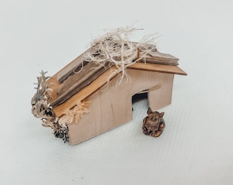 Könnte beinhalten: Ein Miniatur-Holzhaus mit hellbrauner Außenfassade und einem geschichteten Dach. Das Dach besteht aus dünnen, dunkelbraunen Holzlatten. Das Haus ist mit getrocknetem Moos verziert und eine kleine, braune Figur befindet sich vor der Tür.