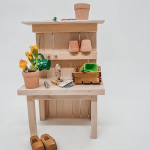 Könnte beinhalten: Ein Miniatur-Gartentisch aus Holz mit einem Terrakotta-Topf, einer grünen Gießkanne und einer kleinen Holzkiste. Auf der Werkbank stehen eine Topfpflanze mit gelben Blüten und weiteres Gartenzubehör. Zwei Holzschuhe stehen am Boden.