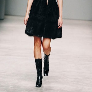 May include: A sleeveless black dress featuring a deep V-neckline, a cinched waist with a thin tie, and a textured, fur-like finish on the skirt. The garment is paired with black, knee-high, lace-up boots with a square toe and a block heel.