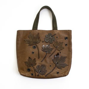 May include: A brown tote bag featuring dark brown leaf-shaped appliqués and black metal grommets scattered across the front. The bag has two olive green fabric handles attached to the top, providing a sturdy grip for carrying personal items.