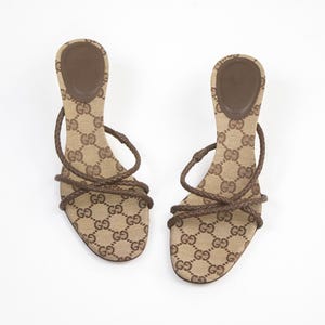 May include: A pair of open-toe sandals featuring a beige canvas footbed with a brown interlocking GG monogram pattern. The footwear includes thin, braided brown leather straps that cross over the forefoot and wrap around the heel area.