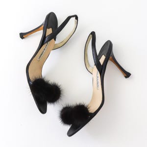 May include: Black leather heeled sandals with a fur pom-pom detail on each strap. The sandals are labeled "Jimmy Choo".