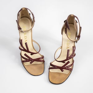 May include: A pair of high-heeled sandals with a brown leather footbed and a burgundy strap design. The sandals have a thin ankle strap and a stiletto heel. The word "PRADA" is visible on the side of the heel.