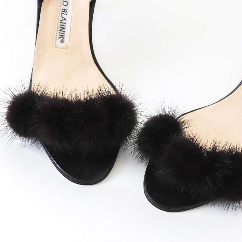 May include: A pair of black heeled sandals with black fur trim around the ankle strap. The sandals have a pointed toe and a thin heel. The brand name "Manolo Blahnik" is visible on the side of the sandal.