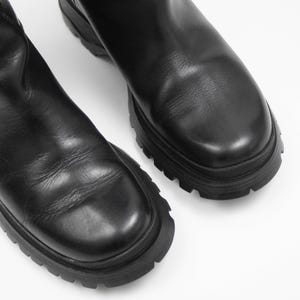 May include: A pair of black leather boots with a chunky, lug sole. The boots have a smooth, matte finish and a rounded toe. The soles are thick and have a deep tread pattern. The boots are set against a white background.