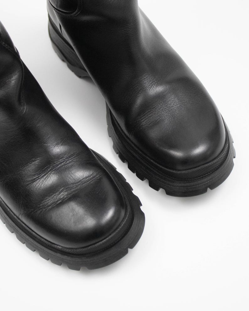 May include: A pair of black leather boots with a chunky, lug sole. The boots have a smooth, matte finish and a rounded toe. The soles are thick and have a deep tread pattern. The boots are set against a white background.