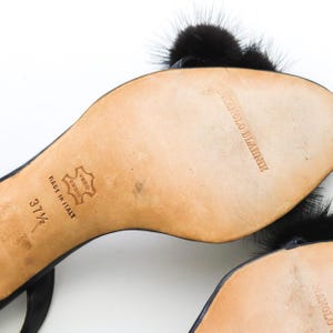 May include: Black leather heeled sandals with black fur pom-poms. The sandals have a buckle closure and the soles are marked "Manolo Blahnik" and "Made in Italy". The size is 37 1/2.