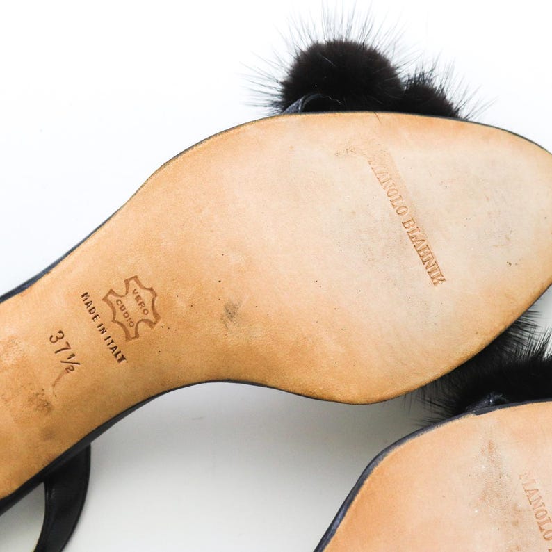 May include: Black leather heeled sandals with black fur pom-poms. The sandals have a buckle closure and the soles are marked "Manolo Blahnik" and "Made in Italy". The size is 37 1/2.