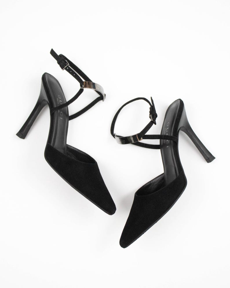 May include: A pair of black high-heeled sandals with pointed toes. The sandals have thin ankle straps with silver-toned metal accents and a buckle closure. The heels are slender and black.