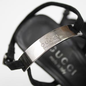 May include: Close-up of a black sandal with a silver metal strap. The strap is embossed with the brand name "GUCCI" and "made in italy". The sandal has a thin ankle strap.