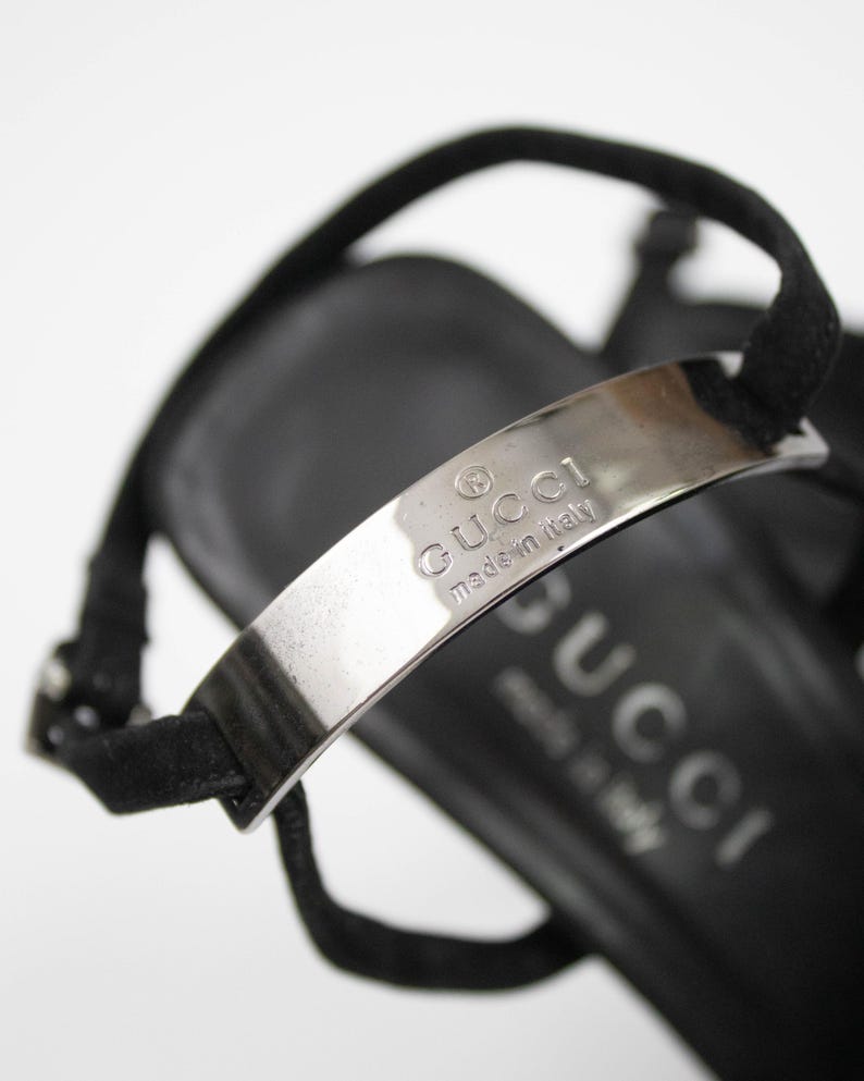 May include: Close-up of a black sandal with a silver metal strap. The strap is embossed with the brand name "GUCCI" and "made in italy". The sandal has a thin ankle strap.
