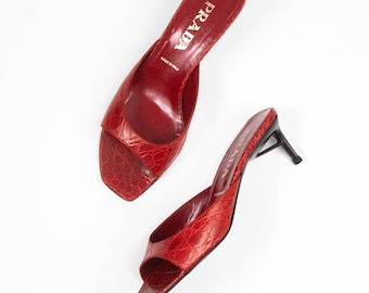 May include: A pair of red crocodile-embossed leather mules with an open-toe design and a black kitten heel. The insole features the white text PRADA and MADE IN ITALY. The shoes have a square toe silhouette and a smooth, polished finish.