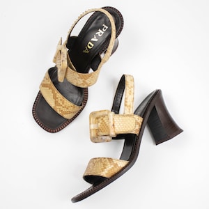 May include: Two Prada sandals with a snakeskin pattern. The sandals have a square toe, a brown sole, and a chunky heel. The straps feature a buckle closure. The word "PRADA" is visible on the insole.