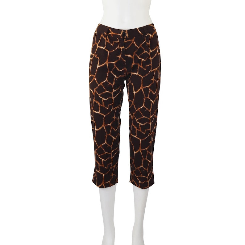 May include: Cropped trousers featuring a brown and tan giraffe print pattern. The trousers are displayed on a white mannequin. The fabric has a geometric design, with dark brown shapes outlined in tan.