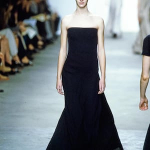 May include: A black strapless gown with a flowing skirt is modeled on a runway. The dress is floor-length and fitted through the bodice. The model has fair skin and red hair. The background shows an audience and other models.