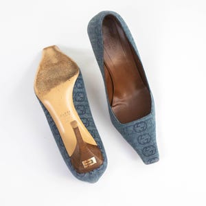 May include: A pair of blue denim pumps featuring a repeating dark blue interlocking G logo pattern. The shoes have a pointed toe, a brown wooden stiletto heel with a gold-tone metal accent, and a brown leather interior lining with the Gucci brand name.