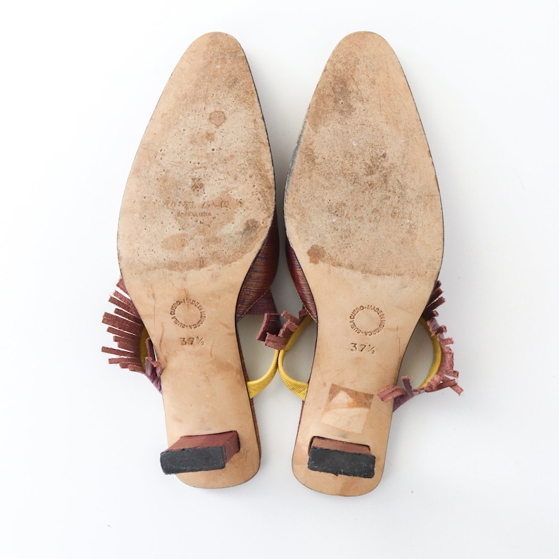 K&ouml;nnte beinhalten: Ein Paar braune Ledersandalen mit Absatz und gelben und burgunderfarbenen Fransenakzenten. Die Sandalen haben eine spitze Zehenpartie und einen kleinen Blockabsatz. Die Gr&ouml;&szlig;e 37 1/2 ist auf den Sohlen aufgedruckt.
