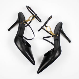 Puede incluir: Un par de zapatos de tacón negros con punta y finas correas en el tobillo. Los zapatos presentan herrajes dorados y un tacón de aguja. Los zapatos se exhiben sobre un fondo blanco.