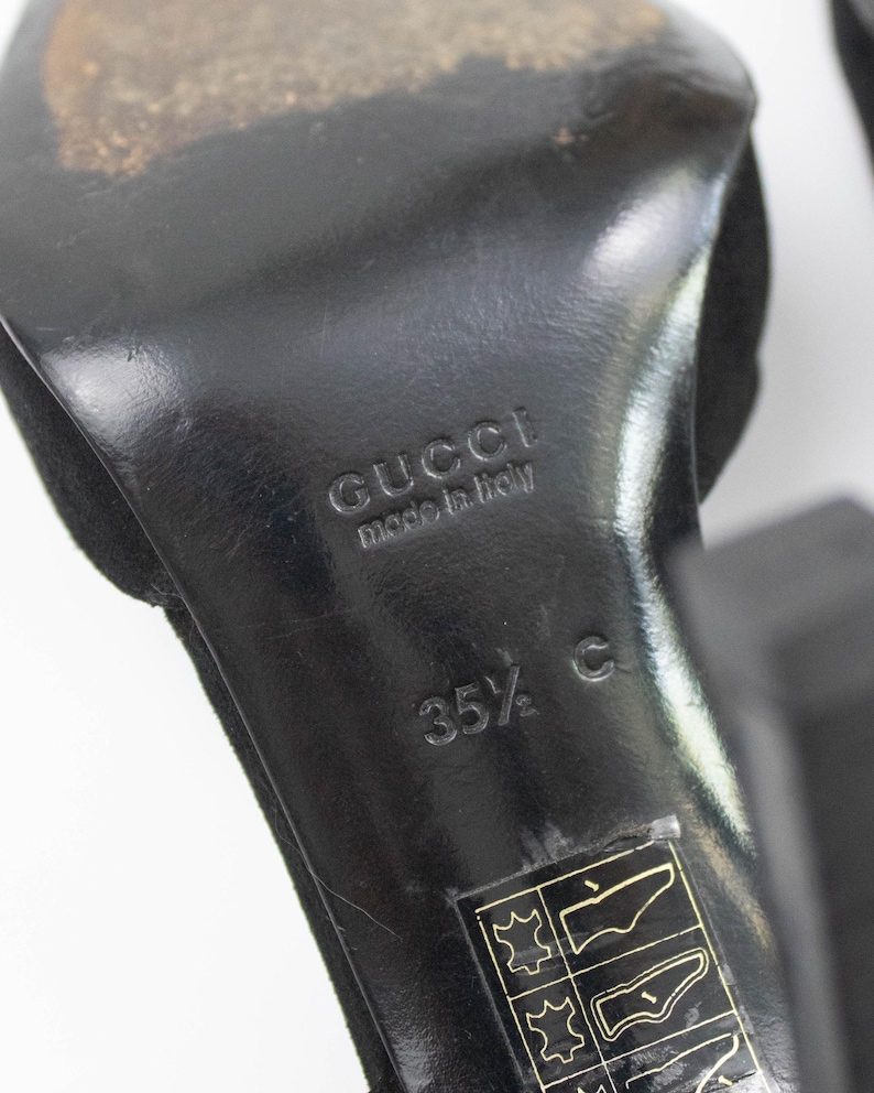 May include: Close-up of a black Gucci high heel shoe sole. The sole is marked with "GUCCI made in italy" and size "35 1/2 C". The shoe has a suede upper.