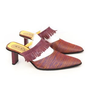 K&ouml;nnte beinhalten: Ein Paar braune und violette Mules mit Absatz und Fransen. Die Schuhe haben eine spitze Zehenpartie und einen wei&szlig;en Ledereinsatz. Der Markenname "MUXA BARCELONA" ist auf der Sohle sichtbar.