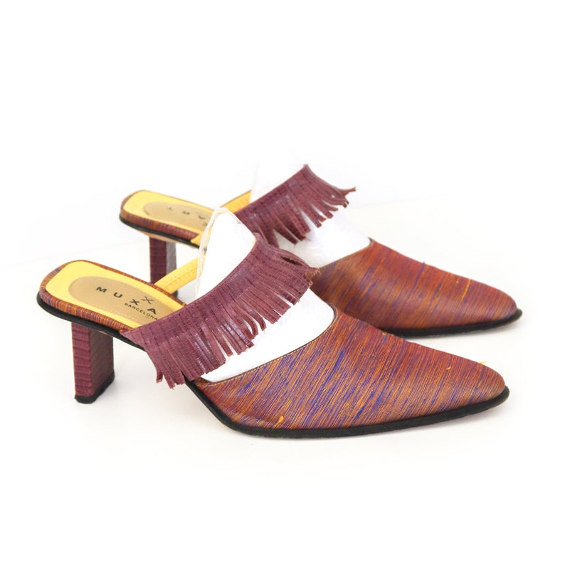 K&ouml;nnte beinhalten: Ein Paar braune und violette Mules mit Absatz und Fransen. Die Schuhe haben eine spitze Zehenpartie und einen wei&szlig;en Ledereinsatz. Der Markenname "MUXA BARCELONA" ist auf der Sohle sichtbar.