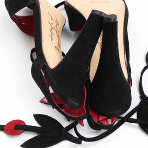 Puede incluir: Sandalias de tac&oacute;n alto de ante negro con detalles en cuero rojo. Las sandalias presentan un dise&ntilde;o de tiras y un elemento floral decorativo. Los tacones son gruesos y las suelas son de color claro.