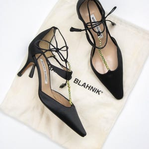 Puede incluir: Un par de zapatos de tac&oacute;n alto de sat&eacute;n negro con puntera afilada y delicadas correas en el tobillo. Los zapatos tienen un tac&oacute;n fino y est&aacute;n adornados con una cadena de cuentas verdes. Descansan sobre una bolsa de tela crema con la palabra "BLAHNIK".