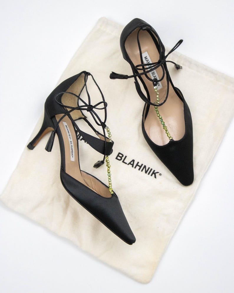 Puede incluir: Un par de zapatos de tac&oacute;n alto de sat&eacute;n negro con puntera afilada y delicadas correas en el tobillo. Los zapatos tienen un tac&oacute;n fino y est&aacute;n adornados con una cadena de cuentas verdes. Descansan sobre una bolsa de tela crema con la palabra "BLAHNIK".