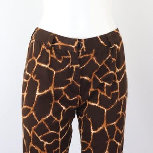 May include: Brown trousers featuring a giraffe print. The trousers have a dark brown base with a pattern of tan and light brown shapes. They have a button closure and belt loops.