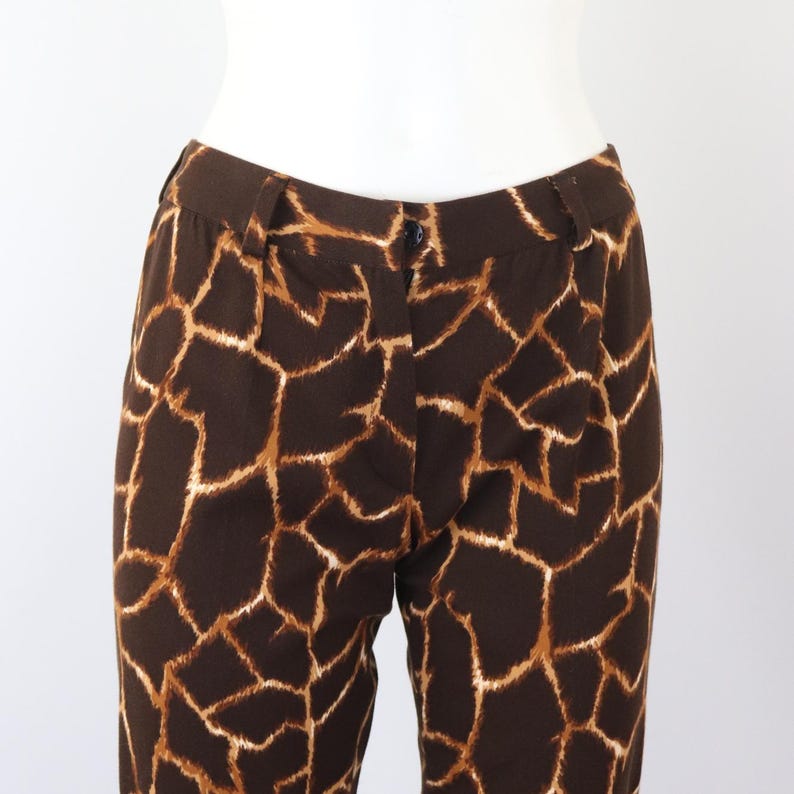 May include: Brown trousers featuring a giraffe print. The trousers have a dark brown base with a pattern of tan and light brown shapes. They have a button closure and belt loops.