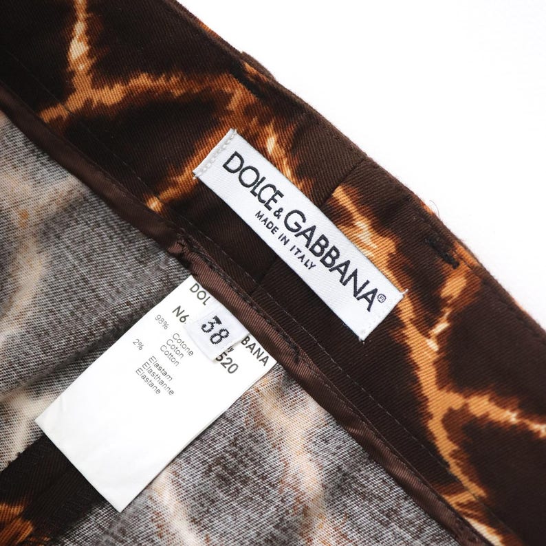 May include: Close-up of brown and tan animal print trousers. The trousers have a dark brown waistband and the tag reads "DOLCE & GABBANA MADE IN ITALY". The tag also lists the size as 38.