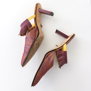 K&ouml;nnte beinhalten: Ein Paar violette und orangefarbene Mules mit Fransenbesatz und gelben Riemen. Die Abs&auml;tze sind blockf&ouml;rmig und die Schuhe haben eine spitze Zehenpartie.