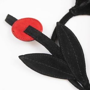 Puede incluir: Una flor de gamuza negra con un acento circular rojo. La flor tiene dos hojas negras y un tallo negro. El acento rojo es un peque&ntilde;o trozo redondo de gamuza con una ranura para que pase el tallo.