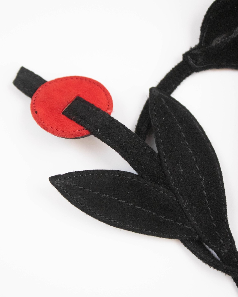 Puede incluir: Una flor de gamuza negra con un acento circular rojo. La flor tiene dos hojas negras y un tallo negro. El acento rojo es un peque&ntilde;o trozo redondo de gamuza con una ranura para que pase el tallo.