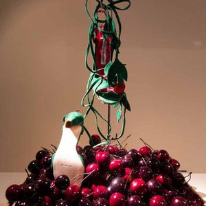 Puede incluir: Una caja de madera rebosante de cerezas frescas y maduras. Un p&aacute;jaro de peluche blanco y verde se encuentra entre la fruta. Una pieza decorativa con una botella roja y hojas y enredaderas de fieltro verde se eleva sobre las cerezas.