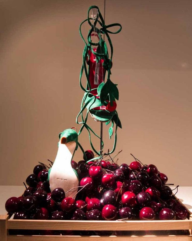 Puede incluir: Una caja de madera rebosante de cerezas frescas y maduras. Un p&aacute;jaro de peluche blanco y verde se encuentra entre la fruta. Una pieza decorativa con una botella roja y hojas y enredaderas de fieltro verde se eleva sobre las cerezas.